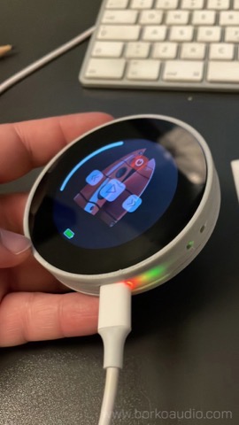 Round wireless remote held in one hand, colour display showing the speaker control screen